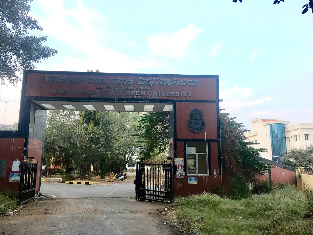 A large brown gate of KSOU, with trees and buildings under a cloudy sky.